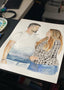 Custom couple portrait being painted in watercolour on a desk, a man and woman embracing beside a paint palette