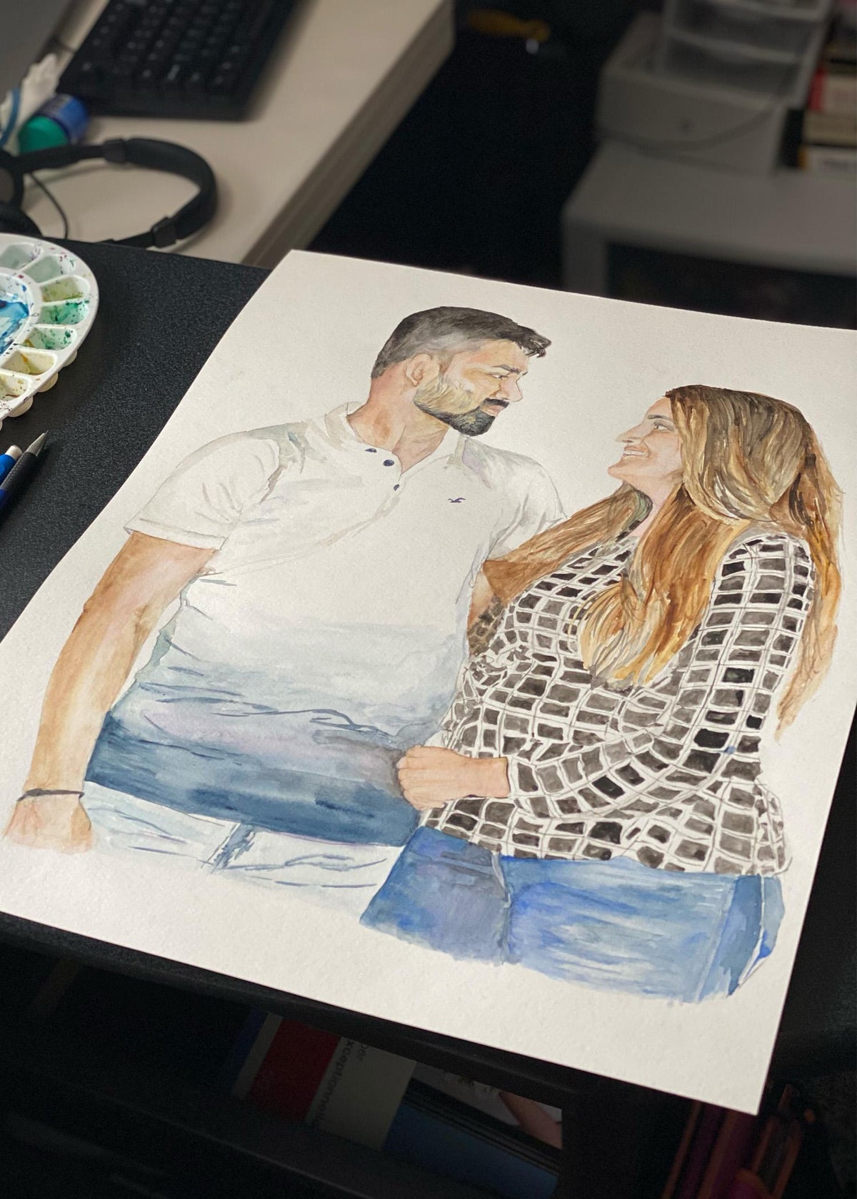Custom couple portrait being painted in watercolour on a desk, a man and woman embracing beside a paint palette Main image