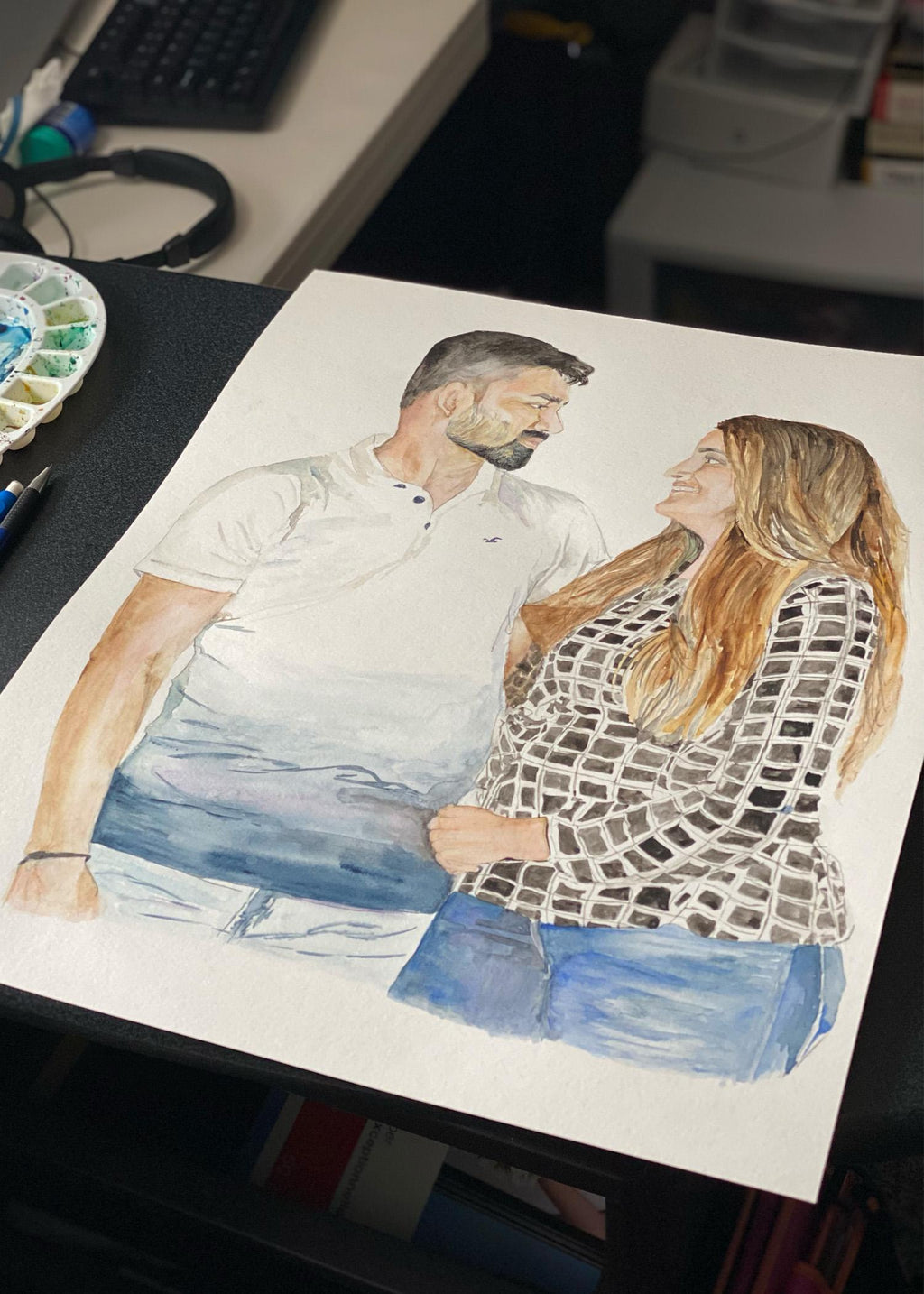 Custom couple portrait being painted in watercolour on a desk, a man and woman embracing beside a paint palette