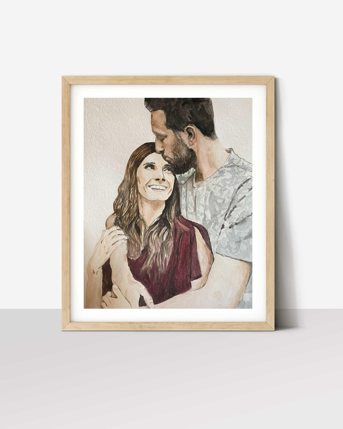 Couple portrait painted from a photo, man kissing woman's forehead, in a white-matted oak frame